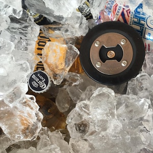 Peut inclure: Un ouvre-bouteille noir avec un dessus en cuivre et les mots "The PuckoOpener" grav&eacute;s sur le dessus. L'ouvre-bouteille est entour&eacute; de gla&ccedil;ons et d'une bouteille de bi&egrave;re Corona Extra.