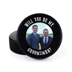 May include: Black hockey puck with a white circle containing a photo of two men in suits. The text "Will you be my groomsman?" is printed around the circle.