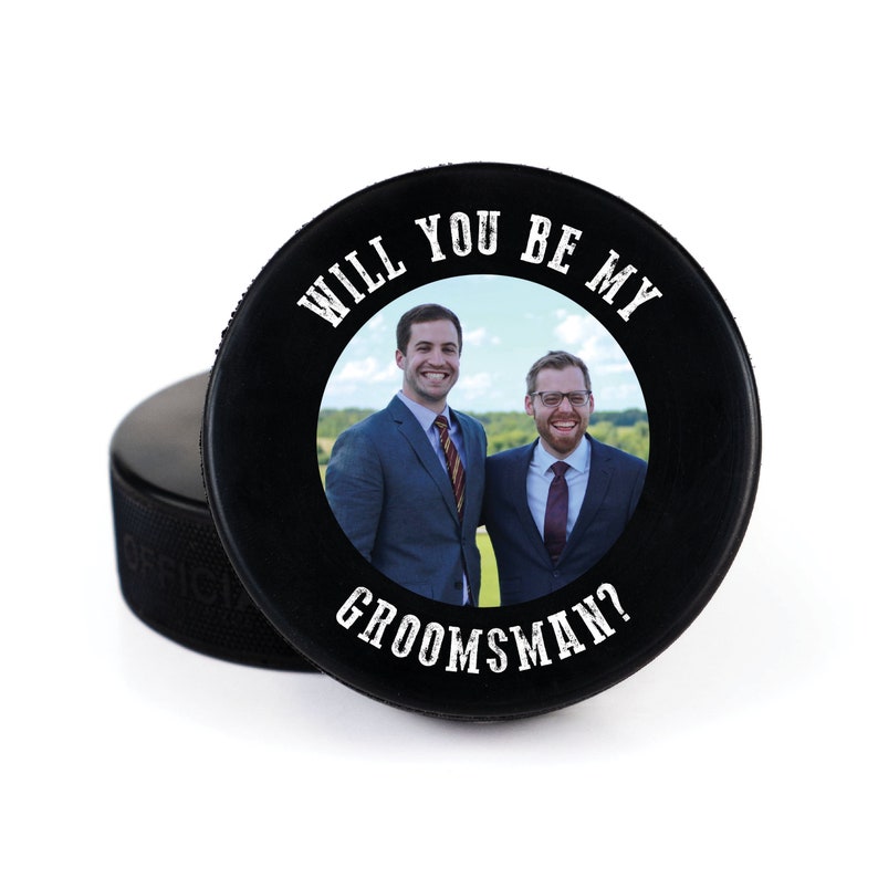 May include: Black hockey puck with a white circle containing a photo of two men in suits. The text "Will you be my groomsman?" is printed around the circle.