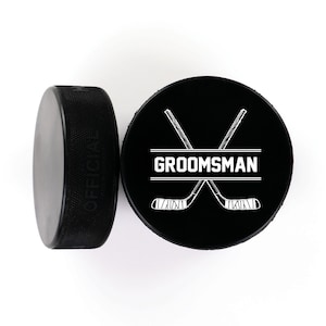 May include: Two black hockey pucks with white designs. One puck has the word "OFFICIAL" printed on the side. The other puck has a pair of crossed hockey sticks with the word "GROOMSMAN" printed in a banner.