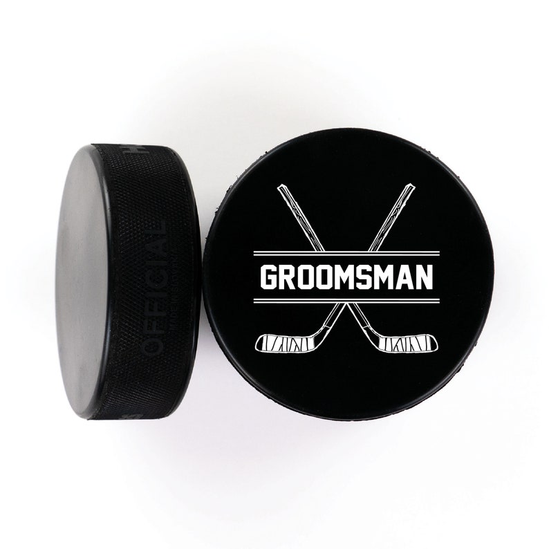 May include: Two black hockey pucks with white designs. One puck has the word "OFFICIAL" printed on the side. The other puck has a pair of crossed hockey sticks with the word "GROOMSMAN" printed in a banner.