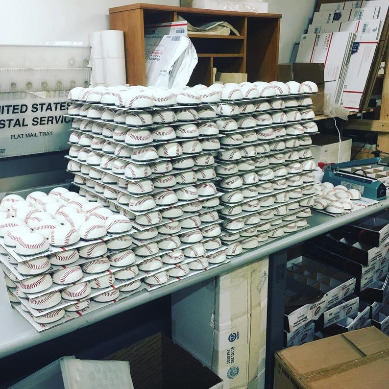 May include: A large stack of white baseballs with red stitching, stacked on top of each other on a table. The baseballs are arranged in rows and columns, and there are many stacks of baseballs in the image.