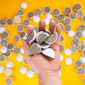 May include: A hand holding a pile of white golf balls on a yellow background. The golf balls are arranged in a random pattern.