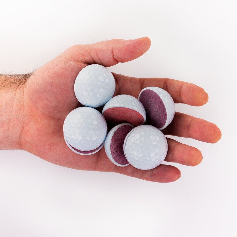 May include: A hand holding a group of six golf ball-shaped candies. The candies are white with a dimpled surface and have a purple filling.