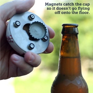 Pu&ograve; includere: Un apribottiglie a forma di palla da baseball con un raccoglitore di tappi magnetico. L'apribottiglie &egrave; in metallo e presenta un design a palla da baseball bianco. Il raccoglitore di tappi &egrave; in metallo e ha una finitura nera. L'apribottiglie viene utilizzato per aprire una bottiglia di birra in vetro marrone.