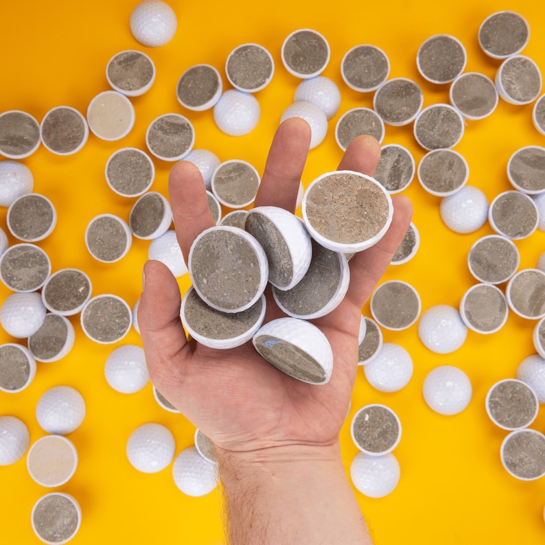 May include: A hand holding a variety of white golf balls, some of which are covered in a brown, textured material. The balls are arranged in a circular pattern on a yellow surface.