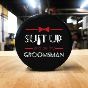 May include: Black hockey puck with white text that reads "Suit Up and be my Groomsman" with a red bow tie and a white tie graphic.