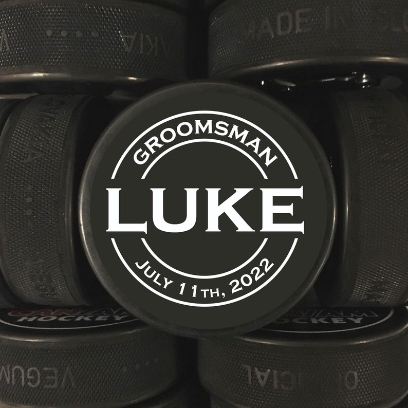 May include: Black hockey puck with a white circle and the text "Groomsman Luke July 11th, 2022".