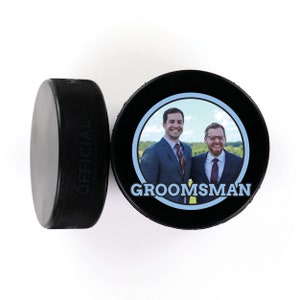 May include: Two men in suits stand together in front of a green field. The man on the left is wearing a blue tie and the man on the right is wearing a red tie. The image is printed on a black hockey puck with the word "GROOMSMAN" in blue letters.