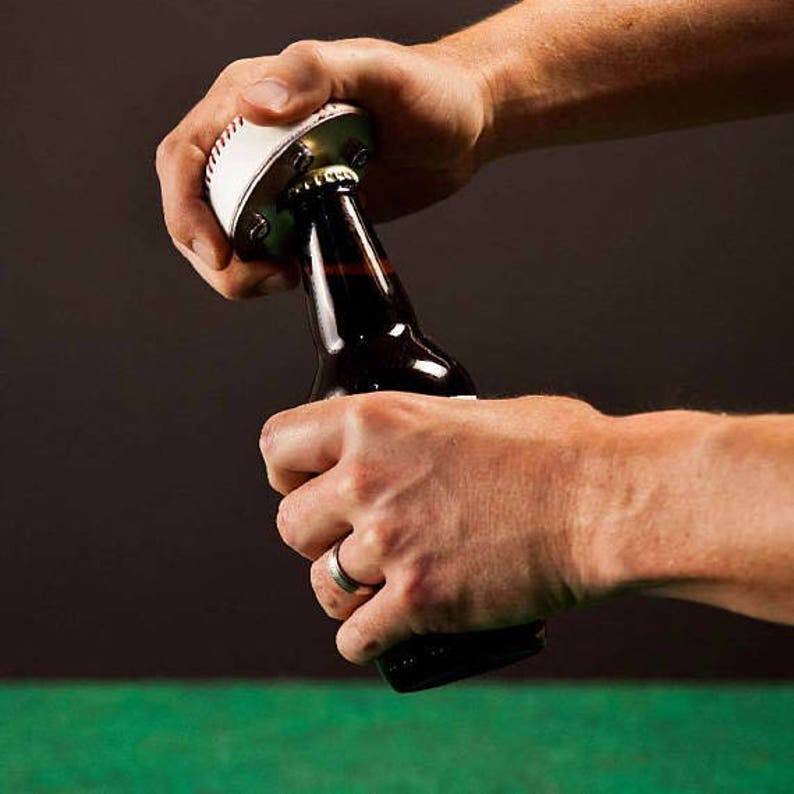 Pu&ograve; includere: Una persona sta usando un apribottiglie a forma di palla da baseball per aprire una bottiglia di birra di vetro marrone.