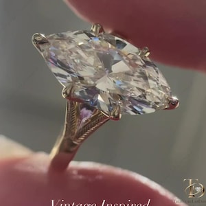 Peut inclure: Une bague en or avec une grande pierre centrale taille marquise fixée par des griffes. L'anneau présente des détails gravés complexes sur les épaules. Le texte "Vintage Inspired" et un logo sont visibles en bas de l'image.