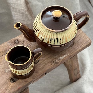 Op de afbeelding: Een bruine keramische theepot met bijpassend melkkannetje, beide voorzien van een crèmekleurige band met verticale goudkleurige druppelpatronen en goudkleurige randen. De theepot heeft een deksel met een goudkleurige knop, handvat en tuit.