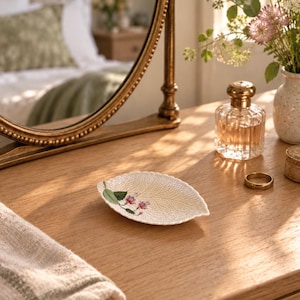 May include: A small, off-white ceramic trinket dish shaped like a leaf with a textured surface and a painted illustration of a green stem with small pink flowers. The dish has a slightly curved, elongated form with a detailed vein pattern.