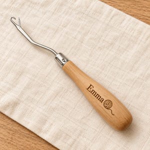 May include: A metal latch hook tool with a bent shaft and a light-colored wooden handle. The handle is engraved with the name Emma and a small graphic of a ball of yarn. The tool features a hinged latch mechanism at the tip.