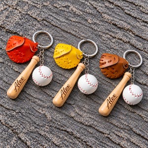 May include: Three keychains featuring a miniature baseball glove in orange, yellow, or brown, a small white baseball, and a wooden bat charm engraved with the name Alex. Each item is attached to a silver-toned metal split ring.