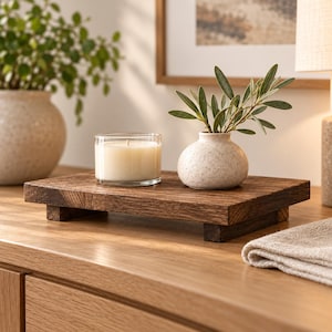 May include: A rectangular dark brown wooden riser with four short feet. A clear glass candle holder containing a white wax candle and a small, speckled, off-white ceramic vase holding green leafy stems sit on the wooden surface.