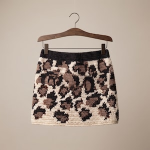 Peut inclure: Une mini-jupe en crochet avec une ceinture noire et un motif léopard répétitif dans des tons crème, marron et noir. Le vêtement est présenté sur un cintre en bois foncé, mettant en valeur la construction en fil texturé et les détails du point.