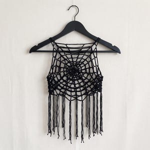 May include: A black crochet crop top featuring a spider web pattern design. The garment has a high neckline, thin shoulder straps, and a straight hemline finished with long, dangling fringe tassels hanging from the bottom edge of the piece.