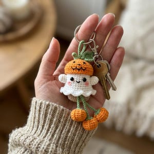 May include: A crochet keychain featuring a white ghost figure wearing an orange pumpkin hat with a jack-o'-lantern face. The ghost has black bead eyes and pink cheeks. Three small orange crochet spheres hang from the bottom on green stems.