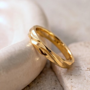 Könnte beinhalten: Ein goldener Ring mit einer polierten, reflektierenden Oberfläche und einer Reihe von subtilen, wellenförmigen Vertiefungen entlang des äußeren Bandes. Das Metall hat einen warmen, hellgelben Ton und eine glatte, runde Form.