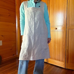 May include: A light beige linen apron with a square neckline and two front pockets. The apron is worn over a light blue zip-up top and blue jeans. The background features wooden doors and paneling.