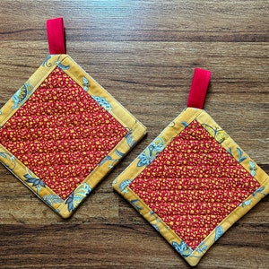 Puede incluir: Dos agarraderas de olla acolchadas rojas y amarillas con un patrón floral y lazos rojos para colgar.