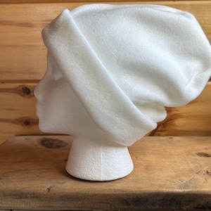 May include: A solid white beanie hat with a thick, folded cuff at the base. The hat is made of a soft, smooth fabric and is displayed on a white foam mannequin head, showing a simple, plain design with no patterns, logos, or additional embellishments.