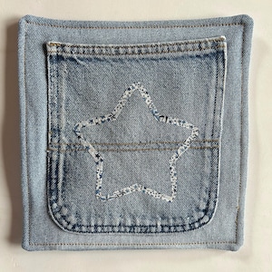 May include: Square denim potholder with a light blue jean pocket sewn on the front. A star shape is appliqued on the pocket with a white and blue floral fabric. The edges are topstitched.