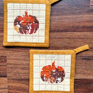 Könnte beinhalten: Zwei gelbe, gesteppte Topflappen mit einem Herbstblatt-Kürbis-Design. Die Topflappen haben eine Schlaufe zum Aufhängen.