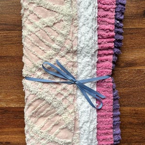 May include: A stack of soft, textured fabric squares in shades of pink, white, and purple, tied with a blue ribbon. The fabrics have a plush, looped texture, creating a cozy and inviting appearance. The stack is neatly arranged, suggesting a set of decorative or functional textiles.