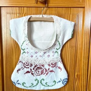 May include: White linen bag with a crocheted trim and embroidered flowers in red, green, and blue. The bag is hanging on a wooden hanger.