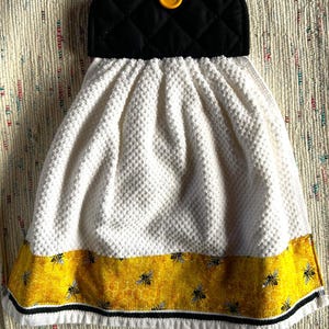 May include: A hanging kitchen towel featuring a white waffle-weave fabric with a yellow border printed with a honeycomb pattern and black bees. The top section is a black quilted fabric flap secured with a single bright yellow button.