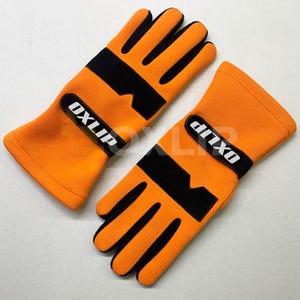 May include: A pair of bright orange gloves with black accents on the fingers and palms. Each glove features a black wrist strap with the white text OXLIP printed on it. The gloves are made of a soft, fleece-like material with a textured finish.