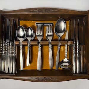 May include: A wooden cutlery tray with carved floral details containing stainless steel flatware, including knives, spoons, forks, and a ladle. A small label in the center reads Stainless by International. The interior is lined with yellow felt.