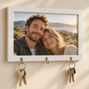 May include: A white rectangular wall-mounted photo frame with a horizontal orientation. The frame features three silver metal hooks attached to the bottom edge, with two sets of metal keys hanging from the outer hooks on the left and right sides.
