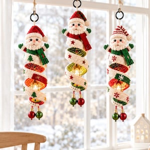 Könnte beinhalten: Drei hängende Spiralornamente mit gehäkelten Weihnachtsmannfiguren an der Spitze. Jedes Ornament hat ein cremefarbenes, spiralförmig nach unten verlaufendes Band, das mit roten und grünen gestickten Sternen und Bäumen verziert ist und mit zwei kleinen glänzenden Kugeln endet.