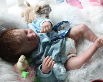 May include: A realistic baby doll with brown hair, wearing a light blue onesie and a diaper, lies on a fluffy white surface. A blue pacifier holder is attached. A stuffed dog and a toy are nearby.