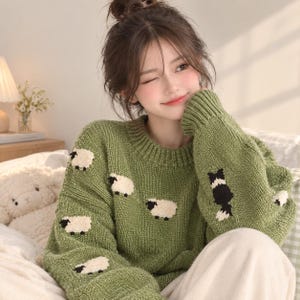 May include: A sage green knit sweater featuring a crew neckline and long sleeves. The sweater is decorated with a repeating pattern of small, cream-colored sheep with black faces and legs, along with a larger black and white sheep motif on the sleeve.