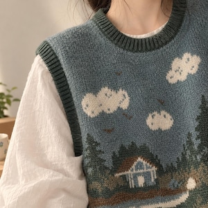 May include: A blue knit sweater vest featuring a landscape pattern with white clouds, dark green pine trees, and a small cabin. The garment has a dark green ribbed crew neckline and armholes, worn over a white long-sleeved shirt with gathered sleeves.