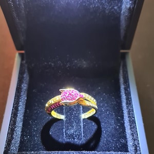 Könnte beinhalten: Ein goldener Ring mit einem zentralen Cluster aus kleinen rosa Steinen in Form einer Blütenknospe, flankiert von Bändern mit einem geometrischen Mäander-Muster. Der Ring liegt in einer schwarz ausgekleideten Box mit LED-Licht.
