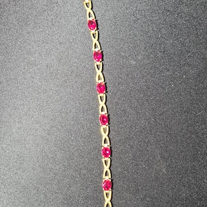 May include: A gold-toned bracelet featuring a series of oval-shaped, vibrant red gemstones set in a repeating X-shaped metal link pattern. The bracelet includes a small chain extender at one end and a lobster claw clasp closure at the other end.