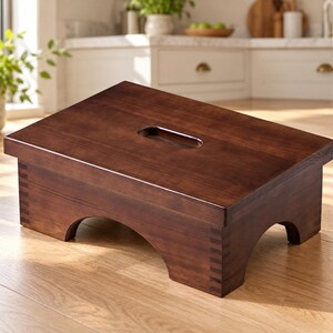 May include: A dark brown wooden step stool with a rectangular top, a centered cutout handle, and arched side supports. The stool features visible finger joint construction at the corners, showcasing a smooth, polished finish throughout the piece.