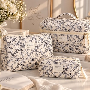 Peut inclure: Un ensemble de trois trousses de toilette matelassées couleur crème avec un motif floral bleu foncé. Chaque trousse est rectangulaire, dotée d'une fermeture éclair avec des tirettes en ruban et d'une étiquette blanche today.mood feel the warm.
