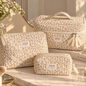 Pode incluir: Um conjunto de três nécessaires acolchoadas em tecido creme com uma pequena estampa floral. Cada bolsa apresenta uma etiqueta retangular branca com o texto today.mood feel the warm. As bolsas possuem zíperes e puxadores de tecido creme.