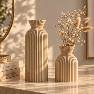 May include: Two beige vases with a vertical ribbed texture and a flared rim. The taller vase is cylindrical, while the shorter vase has a rounded body. The shorter vase contains a bouquet of dried white baby's breath and tan bunny tail grass.