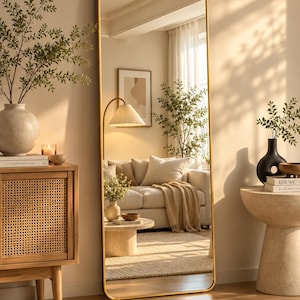 Peut inclure: Un grand miroir rectangulaire sur pied avec des coins arrondis et un cadre fin en métal doré. Le miroir est posé verticalement sur le sol et reflète l'intérieur d'un salon comprenant un canapé, une table basse et un lampadaire à abat-jour plissé.