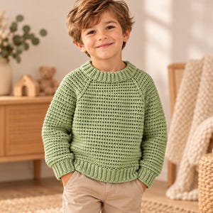 Può includere: Un maglione a maniche lunghe color verde salvia caratterizzato da un motivo a uncinetto testurizzato, un girocollo a coste e polsini a coste. Il maglione è abbinato a pantaloni beige chiaro per un outfit semplice e coerente.