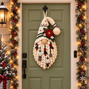 Puede incluir: Un adorno de pared de gnomo tejido a ganchillo con un gorro color crema con rayas verdes y estrellas rojas, una barba grande color crema, una nariz redonda, una flor de pascua roja, hojas verdes, bayas blancas y copos de nieve.