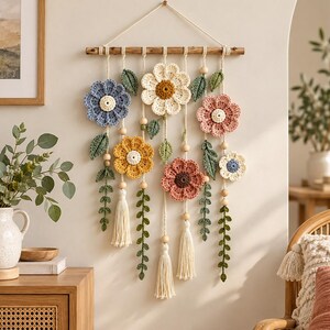 May include: A wall hanging featuring a wooden dowel with hanging crochet flowers in blue, cream, yellow, and pink. The arrangement includes green crocheted leaves, long vine-like stems, natural wooden beads, and cream-colored yarn tassels.