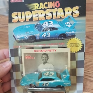 May include: A Racing Superstars toy car featuring a light blue Plymouth with the number 43. The packaging includes the text "Racing Superstars" and "Richard Petty". The toy is designed for ages 5 and up.
